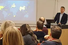 A person giving a talk to a group of people who are seated at a long table. Photo.