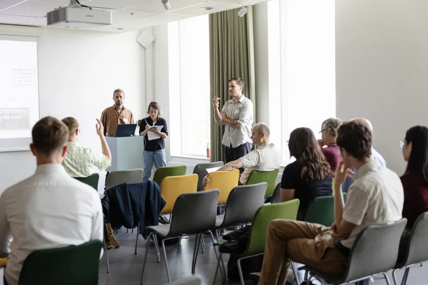 People, some seated and some standing, in a room. Photo.